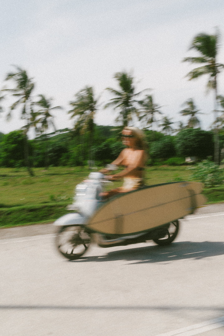Person riding a scooter with a surfboard on a road lined with palm trees wearing Hathaura handmade sterling silver and gold jewellery